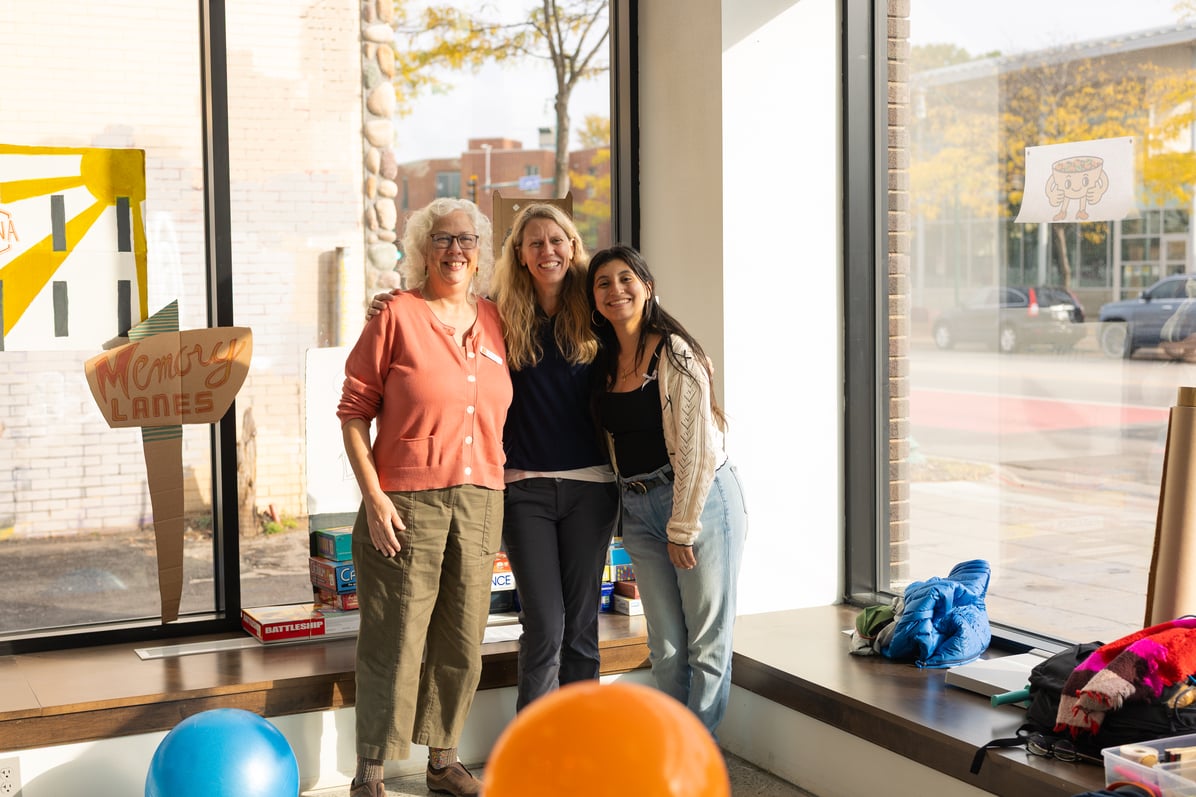 Three people smile in front of sunny windows.